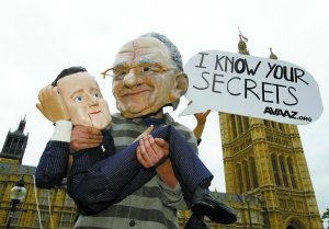 7月13日，一名抗议者扮作默多克，在英国议会大厦前，抱起以英首相卡梅伦为原型的木偶，旁边有一个告示牌，写有：我知道你的秘密。